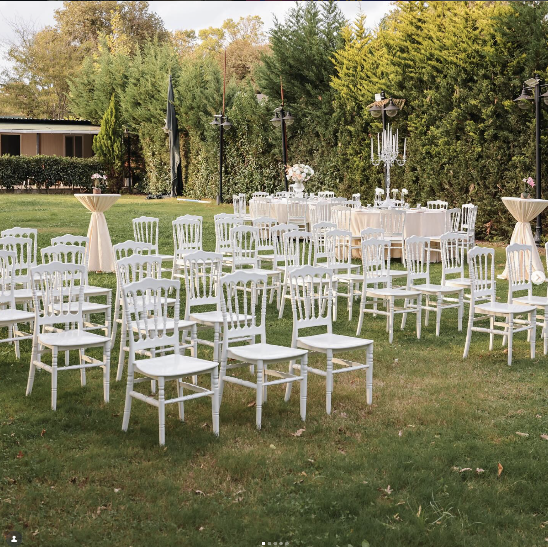 Open-air ceremony aisle with white Napoleon chairs and cream draped tables under evergreens